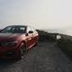 A photo of the front end of an orange BMW 330e sedan in a parking lot near the sea.